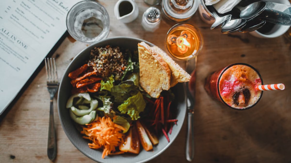 Top view of healthy salad with assorted vegetables and toasted bread placed on wooden table with exotic fresh drink and cutlery